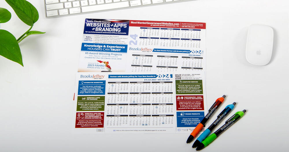 Three 2024 calendars on a table surrounded by pens, a computer mouse, and a keyboard.